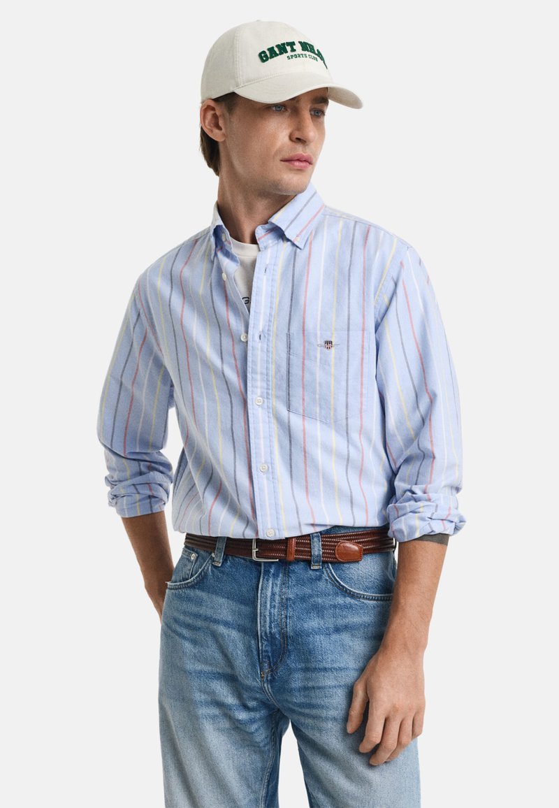 Camisa de botones azul claro con rayas verticales rojas y amarillas, combinada con jeans azul desgastado y una gorra beige con texto verde oscuro.