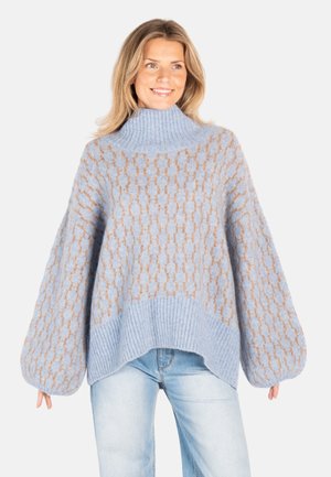 Kvinne iført en løs, lyseblå mønstret genser med høy hals og ballongermer, kombinert med lyseblå jeans, smilende og ser mot høyre.