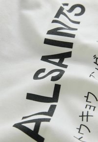 Tissu blanc avec un texte noir audacieux disant « ALL SAINTS » et des caractères supplémentaires, présentant une texture lisse et une légère brillance.