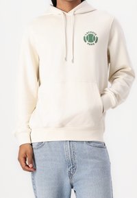 Krémová mikina s kapucí, s přední kapsou, zdobená zeleným logem Lacoste a textem. Vyrobena z měkké tkaniny, má kapuci se stahovací šňůrkou.