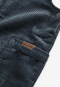 Teren de catifea corduroy albastru închis cu nervuri verticale, având o etichetă din piele maro inscripționată "EST. 1982" și un accent cu un buton negru.