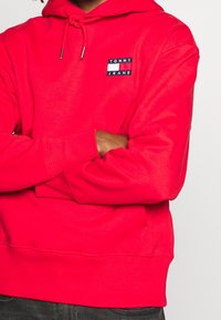 Felpa rossa con cappuccio, tasca frontale a marsupio, cappuccio regolabile con lacci e una piccola etichetta rettangolare con il logo "TOMMY JEANS."