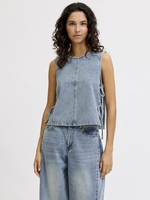 Jeune femme aux cheveux bouclés porte un top en denim bleu clair sans manches avec des liens sur les côtés et un jean large assorti sur fond uni.