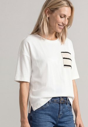 Femme blonde souriante, portant une chemise blanche ample à manches courtes avec une poche tricotée rayée et un jean bleu avec une ceinture.