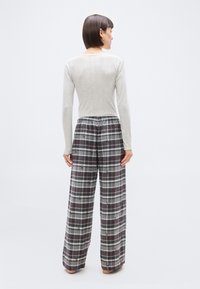 Femme debout pieds nus, tournée de dos, portant un haut à manches longues gris clair et un pantalon large à carreaux gris avec de petits motifs de personnages.