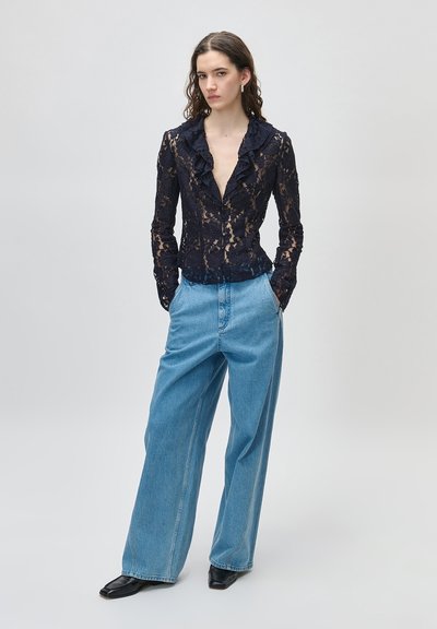 Kobieta stojąca, ubrana w granatową przezroczystą koronkową bluzkę z falbanami, jasnoniebieskie szerokie dżinsy oraz czarne buty i srebrne kolczyki koła.