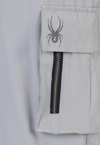 Lichtgrijze stof met een ritszak en marineblauwe accenten, evenals een grijs spinnenlogo op de flap. Gladde textuur met stevige stiksels.
