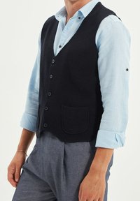 Svart texturerad väst med knappfront och en bröstficka, bärs över en ljusblå rutig skjorta. Har uppvikt ärmar.