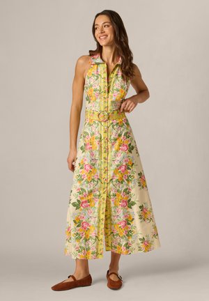 Kvinde smilende, iført en ærmeløs gul blomster maxi kjole med bælte, stående i brune flade sko mod en enkel baggrund.