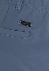 Vaaleansiniset housut, jotka on valmistettu sileästä kankaasta; niissä on musta metallinen logokoriste ja tikattu tasku. Moderni, istuva malli, jossa on joustava vyötärönauha.