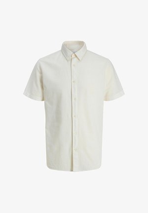 Adolescente con capelli ricci in piedi sorridente, indossa una giacca beige, una camicia bianca con bottoni, pantaloni neri e scarpe da ginnastica bianche, su sfondo semplice.