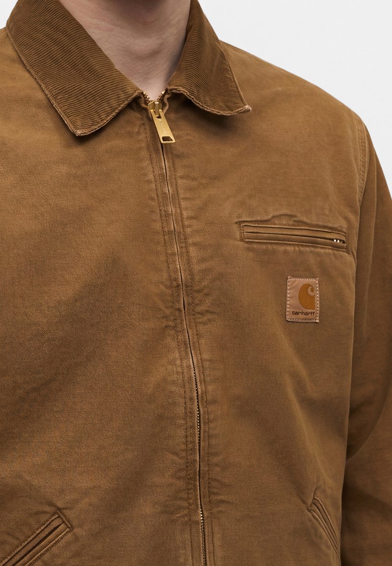 Nahaufnahme einer Person, die eine braune Carhartt-Jacke mit Reißverschluss, Cordkragen und Reißverschlusstasche auf der Brust trägt.