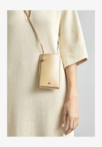 Tan leren telefoonhouder met een gladde textuur en een gouden logo, opgehangen aan een band, gepresenteerd tegen een lichtbeige gebreid jurk.