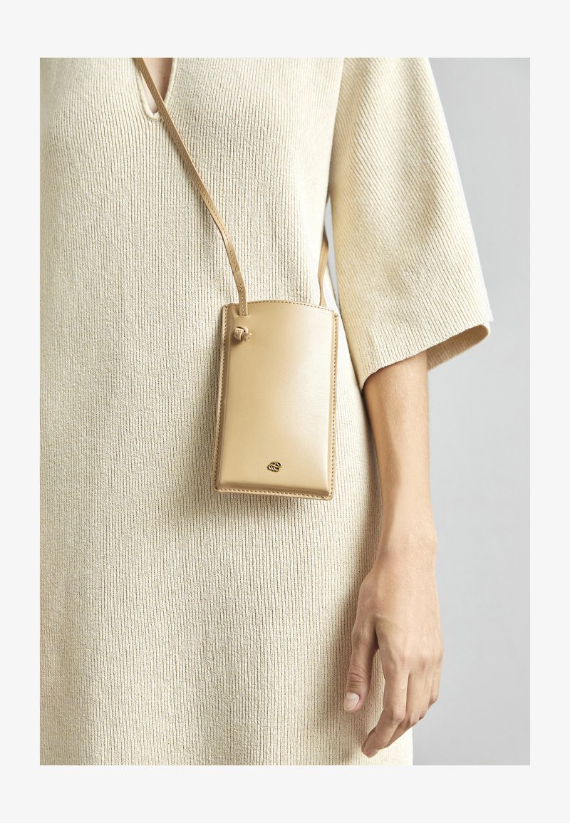 Τηλεφωνική θήκη από καφέ δέρμα με λείο υφή και χρυσό λογότυπο, κρεμασμένη από ένα λουρί, παρουσιασμένη πάνω σε ένα ανοιχτό μπεζ πλεκτό φόρεμα.