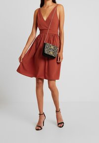 Šatník bez rukávů v rezavé barvě s véčkovým výstřihem a nařaseným pasem, doplněný černou vzorovanou kabelkou a černými sandály na vysokém podpatku.