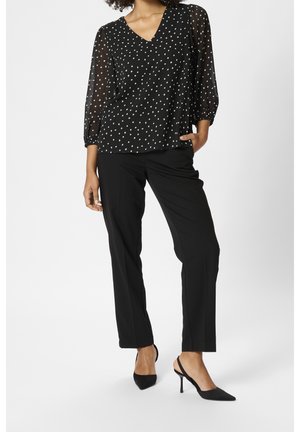 Blouse à pois noirs avec manches transparentes, col en V, portée avec un pantalon noir et des escarpins à bout pointu. Texture lisse et silhouette élégante.