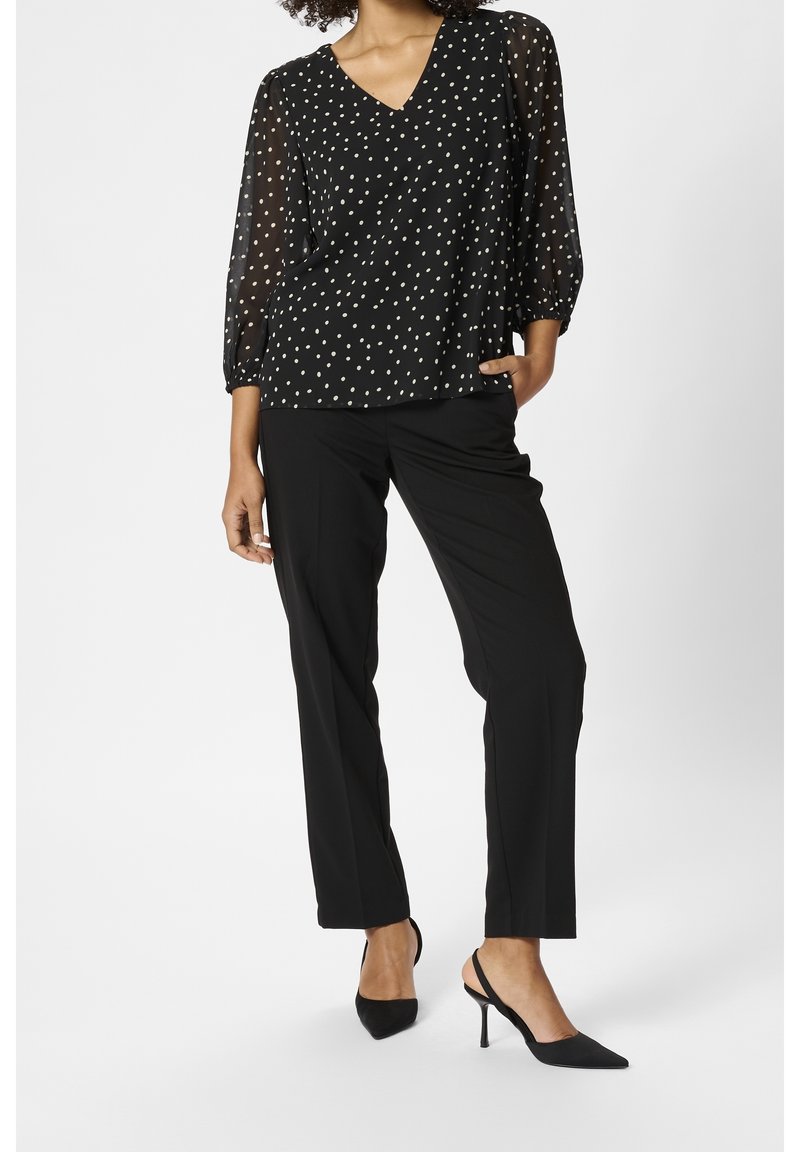 Blouse à pois noirs avec manches transparentes, col en V, portée avec un pantalon noir et des escarpins à bout pointu. Texture lisse et silhouette élégante.