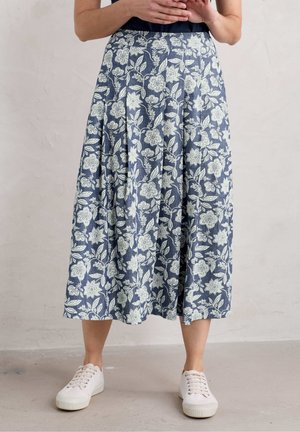 Kvinde iført en blå midi-nederdel med hvide blomsterprint og hvide sneakers, stående mod en ensfarvet lys væg.
