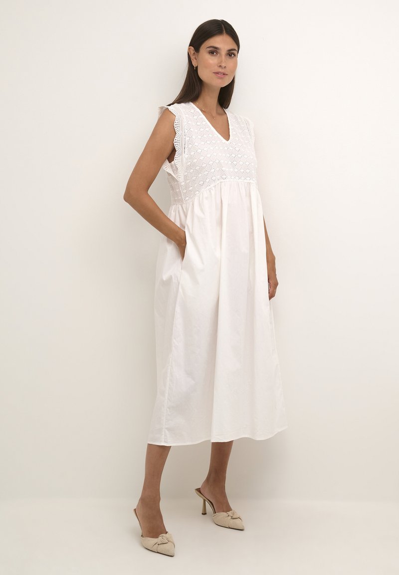 Bílá bavlněná šaty s výstřihem do V, krajkovými detaily na ramenou a rozšířenou siluetou s kapsami. Hladká textura, délka ke kolenům.