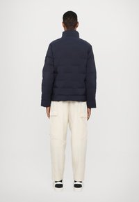 Jachetă puffer bleumarin cu guler înalt, prezentând un design matlasat. Pur tată deasupra pantalonilor cargo de culoare crem. Textură netedă și croială confortabilă.