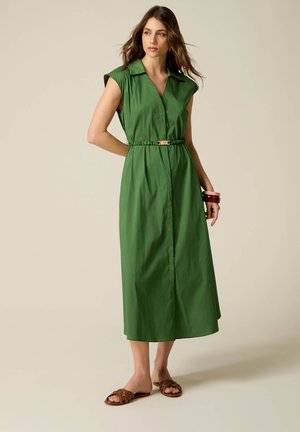 Donna con abito midi verde senza maniche con cintura e sandali marroni a ciabatta, in piedi davanti a uno sfondo beige uniforme.
