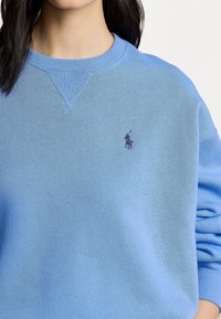 Ljusblå sweatshirt med ribbat mönster, rundad hals, och en liten marinblå logotyp på vänster bröst. Har lång ärm och en avslappnad passform.