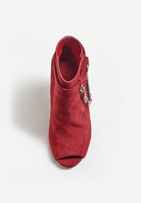 Bottine à talon ouverte en daim rouge avec un détail en chaîne argentée sur le côté extérieur, vue de dessus sur fond blanc.