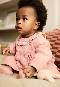 Vestito di mussola rosa con ricami floreali, collo arricciato e maniche lunghe. Bambino seduto tiene un peluche, con sfondo testurizzato.