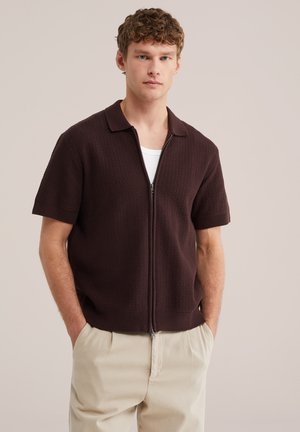 Młody mężczyzna z kręconymi włosami, ubrany w ciemnobrązowy sweter na zamek z krótkim rękawem, nałożony na białą koszulę i beżowe spodnie, stoi z rękami w kieszeniach.