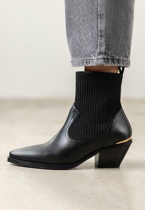 Botte de cheville en cuir noir avec tige en tricot côtelé, bout pointu et un talon épais avec un accent doré. Portées avec un pantalon gris court.