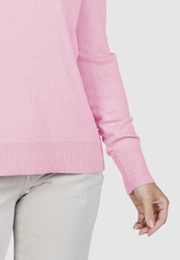 Persoon in een lichtroze trui met lange mouwen en lichtgrijze broek, met de rechterhand ontspannen naast zich.