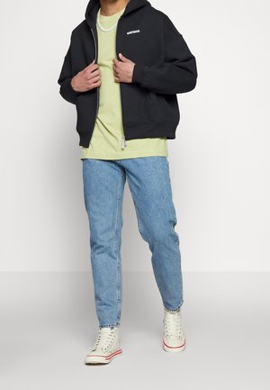 Personne portant un t-shirt vert clair, un sweat à capuche noir zippé, un jean bleu, des baskets montantes blanches et un collier chaîne argenté sur fond blanc.