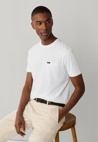 Hombre sentado en un taburete de madera, vistiendo una camiseta blanca de manga corta con bolsillo en el pecho y pantalones beige con un cinturón oscuro.