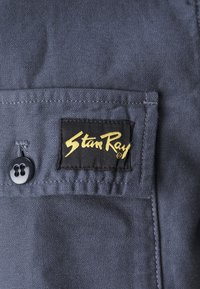 Dark blue fabric with a textured surface featuring a black label with gold lettering. Includes a round black button.