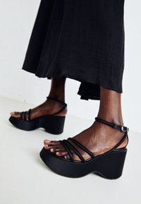 Black wedge sandals featuring multiple thin straps, adjustable ankle buckle, textured surface, and a rounded heel shape.