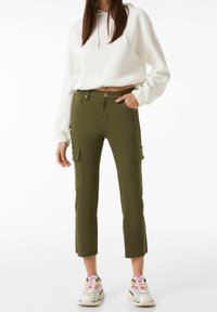 Žena v olivově zelených zkrácených cargo kalhotách, bílé zkrácené mikině a vícebarevných sneakers stojí s jednou rukou v kapse.