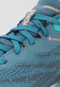 Gros plan sur une chaussure de course bleue Hoka Speedgoat avec des lacets détaillés et une étiquette Gore-Tex sur une tige en tissu texturé.