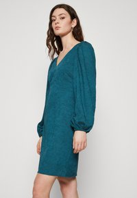 Robe courte teal en tissu texturé, décolleté en V et manches longues bouffantes. Le design présente un corsage ajusté et une jupe évasée.