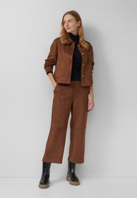 Braune Wildlederjacke mit Knöpfen, kombiniert mit passenden braunen Wildleder-Breitehosen. Ein schwarzer Rollkragenpullover und schwarze Stiefeletten runden das Outfit ab.