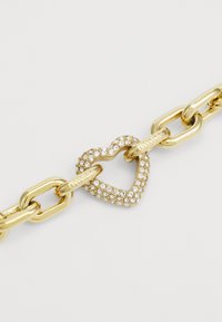 Bracciale in catena dorata con un collante a forma di cuore, caratterizzato da piccoli cristalli e inciso "GUESS," con sezioni lisce e lucide.