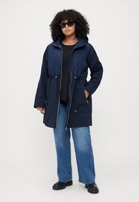 Marineblå hettejakke med quiltet kropp, snor i midjen og lommer, sammen med lyseblå vide jeans og svarte støvler.
