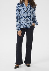 Ljusblå blus med ett geometriskt mönster i marinblått och guldaccenter, kombinerad med mörka utsvängda jeans och svarta klackskor.