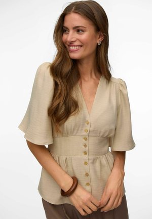 Smilende kvinne med langt brunt hår som har på seg en beige bluse med knapper og vide ermer, samt et trearmbånd på venstre håndledd, ser mot høyre.