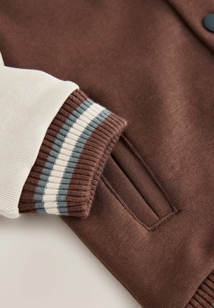 Bruine sweatshirtstof met een contrasterende roomkleurige mouw. Ribbels aan de manchetten hebben blauwe en witte strepen. Inclusief een zijzakdetail.