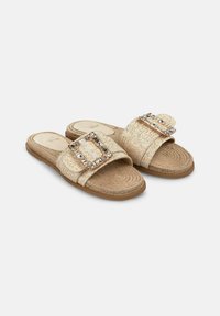 Gold woven slide sandals featuring a wide strap with a rectangular rhinestone embellishment, textured insole, and flat, natural color sole.