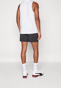 Vit ärmlös skjorta, svarta shorts, vita strumpor och svarta/röda träningsskor. Synliga muskulösa armar och ben från en bakåtvänd perspektiv.