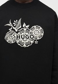 Μαύρο φούτερ με λευκά κεντημένα πουλιά και λουλούδια που περιβάλλουν τη λέξη «HUGO» στο κέντρο του στήθους.
