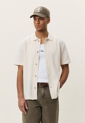 Junger Mann trägt ein beiges Kurzarmhemd über einem weißen Tanktop, einen braunen geflochtenen Gürtel, braune Jeans und eine graue Kappe mit Globus-Design.