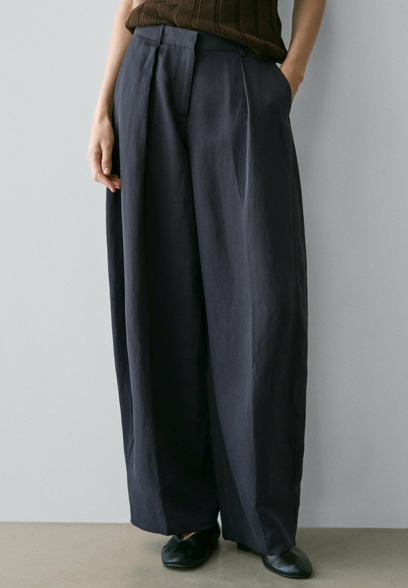 Personne portant un pantalon large sombre, un haut en maille marron sans manches et des chaussures noires, debout contre un mur gris clair uni.