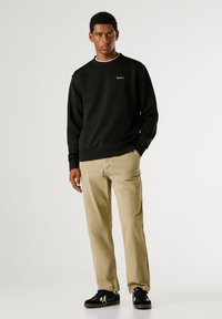 Sweatshirt preto com punhos e bainha canelados, apresentando um pequeno logotipo. Combinado com calças de veludo cotelê bege e ténis pretos.